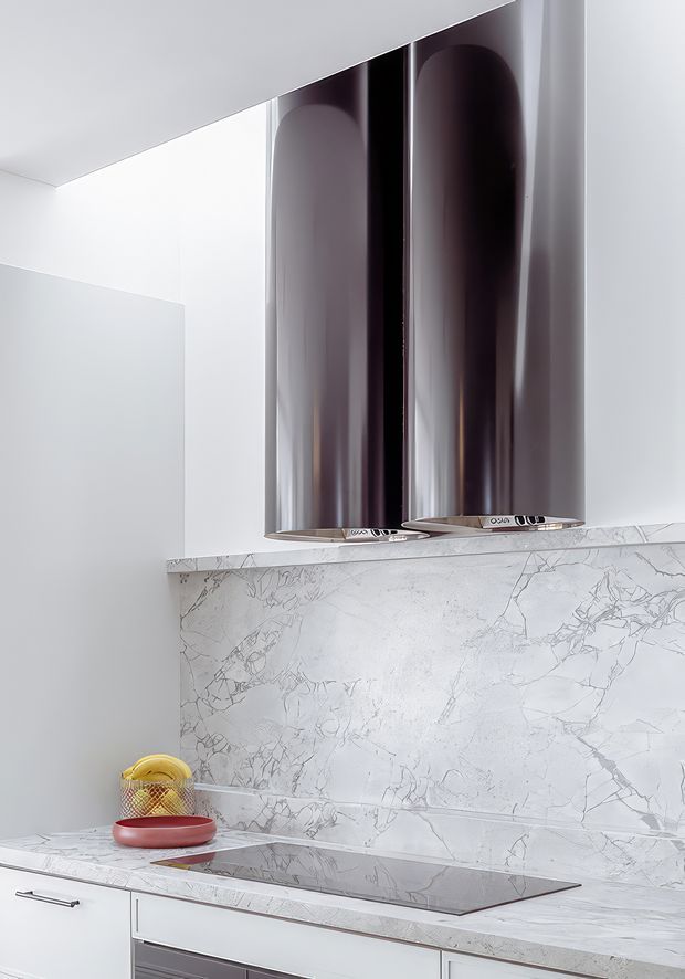 A minimalist contemporary kitchen vignette featuring a white marble or stone benchtop and full-height splashback with prominent grey veining, a dark glass induction cooktop, white cabinetry, and a large sculptural polished metal rangehood. The space is bright, monochromatic, and highly refined.