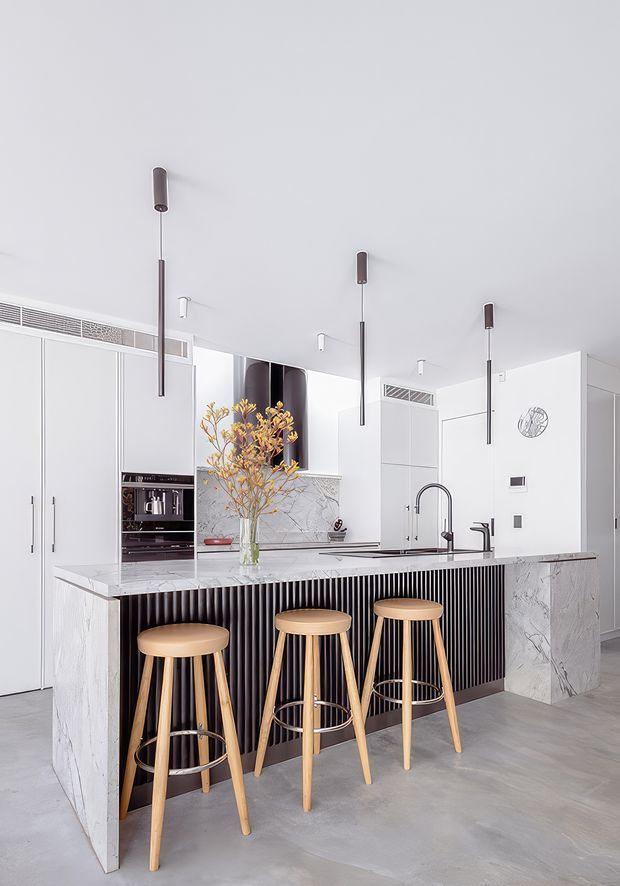 Bright contemporary kitchen featuring a large marble-look island with a black vertical-ribbed base, integrated sink and tapware, white full-height cabinetry, and a built-in coffee appliance. Three light timber bar stools sit at the island, while slim pendant lights hang from the white ceiling. The space has a clean, refined monochromatic palette with warm timber accents and polished stone surfaces.