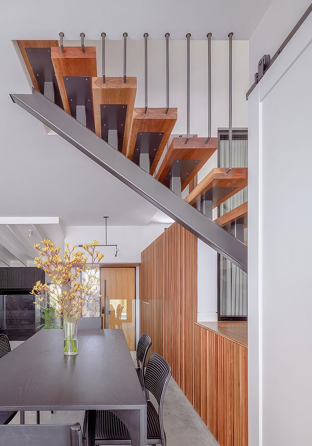 A bright contemporary dining and living interior featuring a dramatic suspended timber staircase with dark metal supports and a steel diagonal balustrade. The room includes a long dark dining table with black wire-frame chairs, warm vertical timber wall panelling, white walls and ceiling, and glimpses of a kitchen or living area beyond.