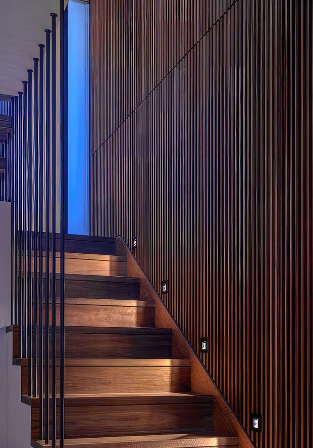 A dramatic interior stairway with warm timber treads and risers, a slim dark metal balustrade, and a tall wall clad in closely spaced vertical timber battens. Small recessed step lights illuminate the staircase, while a vivid blue-lit vertical opening creates a cool contrast. The composition emphasizes the stair circulation and textured feature wall.