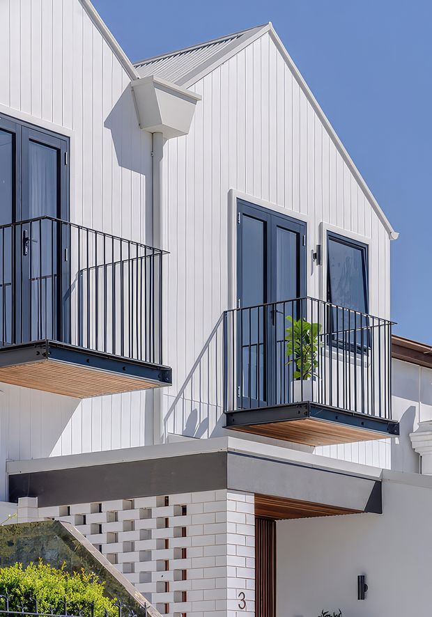 Exterior architectural photograph of a contemporary multi-level residential building with white vertical cladding, steep gabled roof forms, dark-framed doors and windows, and projecting balconies with slim black metal balustrades. Timber soffits and a patterned white-and-grey masonry screen add contrast. Bright clear daylight emphasizes the crisp coastal-inspired facade.
