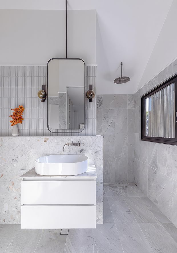 Bright contemporary bathroom featuring a floating white vanity with a round vessel basin, pale terrazzo or stone surfaces, vertically lined wall tiles, large-format grey patterned tiles in the shower and floor, a black-framed window, round wall sconces, and a rounded rectangular mirror. The room has a clean, light, highly coordinated finish palette.