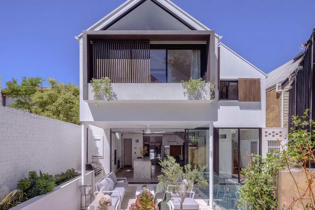 Front exterior view of a contemporary multi-storey residence with a steep gable roof, white rendered walls, dark-framed glazing, and a projecting upper-level balcony. The ground floor opens onto a landscaped courtyard or terrace with outdoor seating, while vertical timber screening and planted balcony edges add privacy and texture.