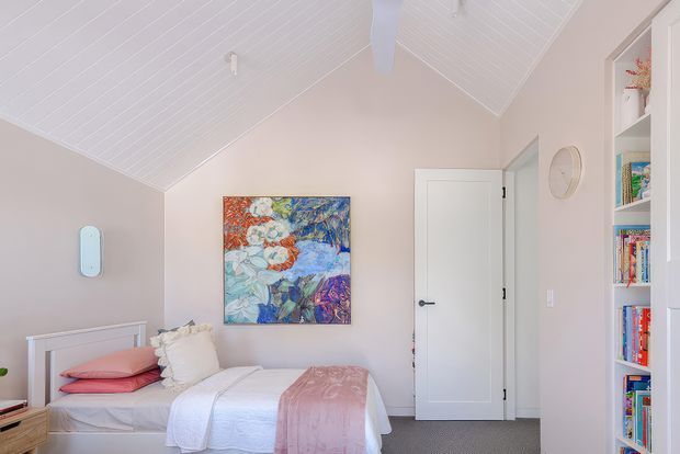Bright, compact bedroom with a single bed dressed in white and blush pink bedding, pale blush walls, a white vaulted V-groove ceiling, and built-in white shelving filled with books. A colourful floral artwork provides the main visual accent, while a white door and soft carpet complete the room.