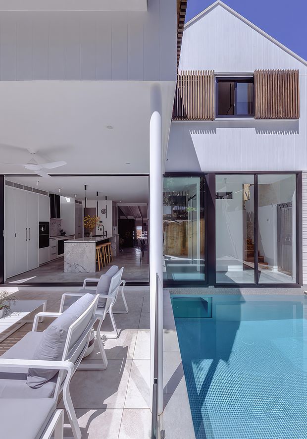 A bright contemporary residential courtyard viewed from the pool terrace, showing an open-plan kitchen and living area on the left and a two-storey white-clad wing on the right. Large glazed doors open toward a blue swimming pool, while the upper level features a pitched gable roof, vertical white cladding, and timber screening around a balcony or window. White outdoor loungers and a slim structural column frame the scene.