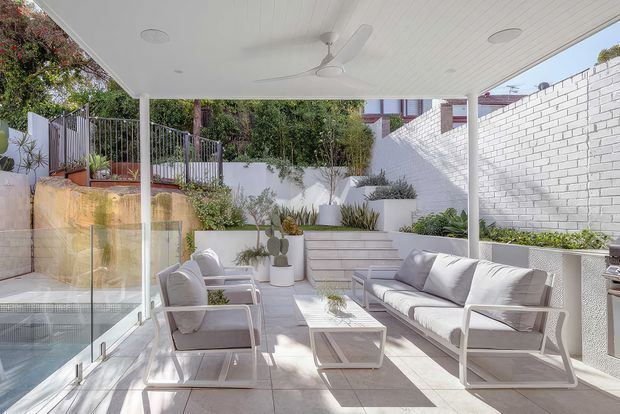 Bright, contemporary covered outdoor lounge with a white-lined ceiling, ceiling fan, glass balustrade, pale tiled floor, white outdoor sofa and armchairs, and a raised landscaped garden bordered by white masonry walls.