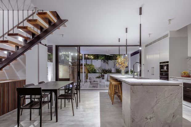 A bright, open-plan contemporary kitchen and dining area with a dramatic floating timber-and-metal staircase. The kitchen features a large veined stone island with bar stools, streamlined white cabinetry, integrated appliances and a dark-framed glazed wall opening to a landscaped courtyard. A dark dining table and chairs sit beneath the stair, while slim pendant lights and pale flooring create a clean, modern atmosphere.