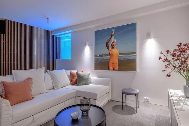 A compact, contemporary living room with a large white sectional sofa, pastel accent cushions, a round black coffee table, and a small upholstered side table. White walls are illuminated by minimalist wall sconces, while a vertical timber slat feature wall and blue-lit recessed niche add visual interest. A large beach photograph serves as wall art, and a white console with a vase of branches sits to the right.