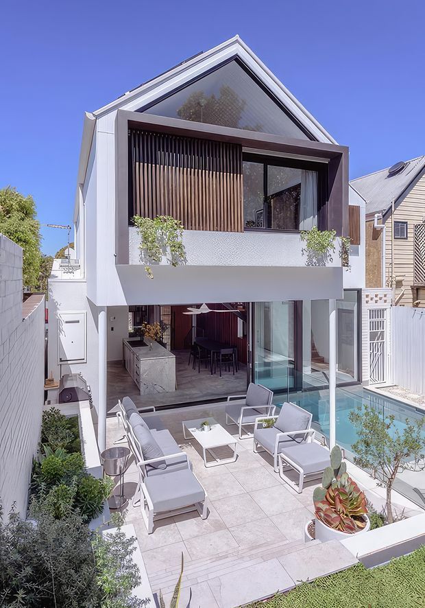 A bright, narrow urban residence viewed from the rear garden, featuring a contemporary multi-level facade with a steep gabled roof, projecting dark-framed upper balcony, vertical timber screening, white rendered walls, and extensive glazing. The ground level opens to a covered outdoor entertaining area with an outdoor kitchen, dining table, lounge seating, and a tiled terrace beside a narrow rectangular swimming pool. Landscaped planting borders the terrace, with neighboring homes visible on either side.