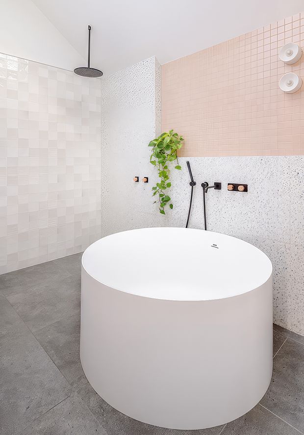 Bright contemporary bathroom featuring a large freestanding white oval bathtub, walk-in shower with a black ceiling-mounted rain shower, wall-mounted fixtures, grey floor tiles, pale pink small-format tiles, white patterned terrazzo-like wall surfaces, and a trailing green plant.