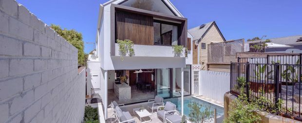 Exterior view of a contemporary multi-storey residence with a white rendered facade, prominent upper-level timber privacy screening, large glazed openings, and a compact backyard pool and outdoor entertaining area. The property is situated in a dense suburban setting with neighbouring homes visible.