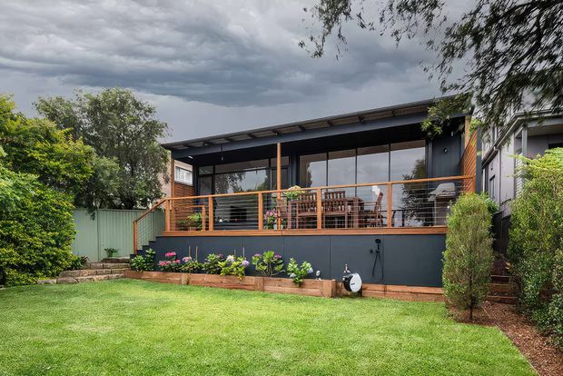 A contemporary single-storey backyard extension or pavilion with dark exterior cladding, broad glazed openings, a shallow skillion roof, and a raised timber deck with orange-toned balustrading. The structure overlooks a manicured lawn and planted garden beds, with outdoor dining furniture visible behind the glazing and under the covered deck.