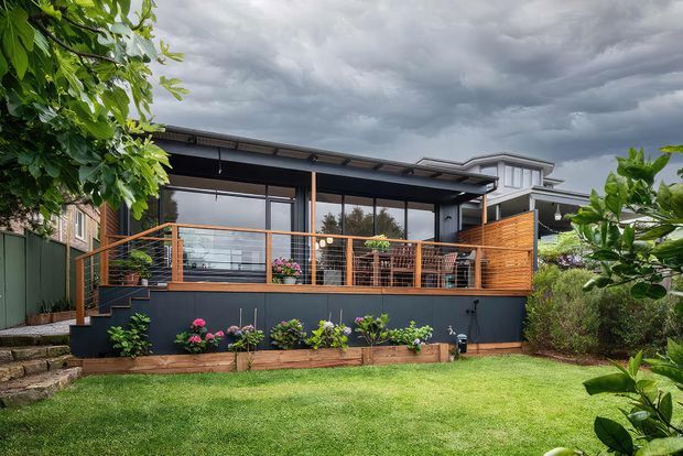 Exterior view of a contemporary single-storey home extension or outdoor living area, featuring a dark metal-clad structure with extensive glazing, a covered terrace, timber privacy screens, horizontal wire balustrades, and a landscaped lawn with raised garden beds. The property sits on a gently sloped suburban site beneath an overcast sky.