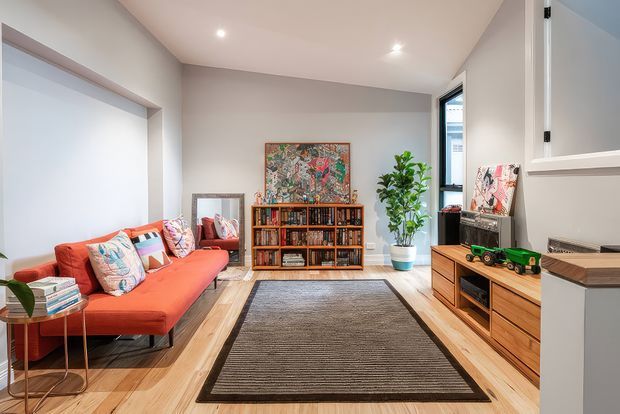 A bright, warm living room with a vivid coral-red sectional sofa, patterned cushions, timber flooring, a large textured area rug, and extensive mid-tone timber cabinetry and shelving. The room includes a bookcase filled with books, framed artwork, indoor plants, a low media console, and a partially visible kitchen or adjoining joinery area on the right.