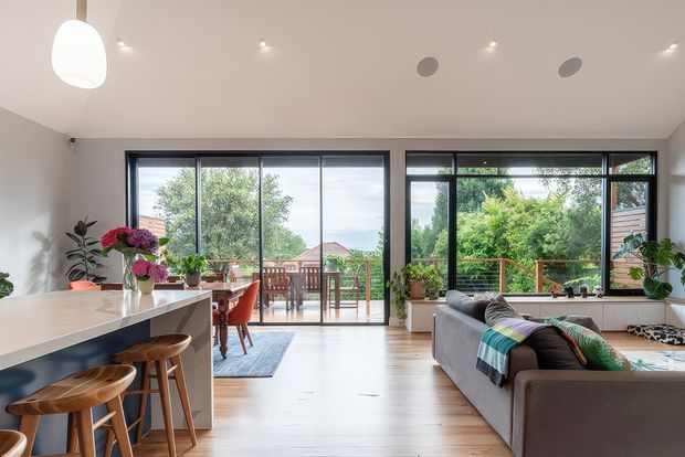 Bright open-plan living space with a contemporary kitchen island in the foreground, timber stools, dining area, and a grey sectional sofa. Large black-framed glazed doors and windows open toward a timber deck and leafy suburban garden. The room has pale walls, light timber flooring, recessed ceiling lights, a sculptural pendant, and colourful floral and textile accents.