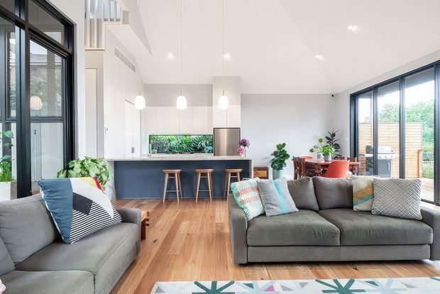 Bright open-plan contemporary living area with a large grey sectional sofa and matching armchair in the foreground, timber flooring, and a compact kitchen with a dark island and bar stools behind. Full-height black-framed glazing opens toward a planted outdoor area, while pendant lights, indoor plants, and patterned cushions add colour and texture.