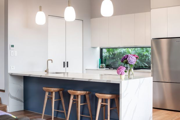 Bright contemporary kitchen with a large island featuring a dark blue-grey front, pale stone benchtop, and seating for three on timber stools. White flat-panel cabinetry, integrated appliances, pendant lights, timber flooring, and a garden-facing window create a clean, refined interior.
