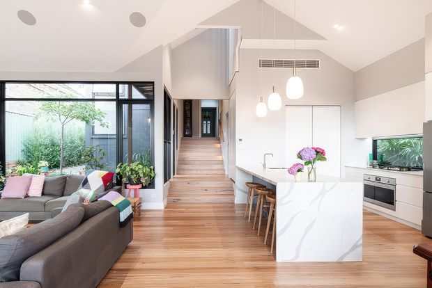 Bright open-plan contemporary interior combining a living room, kitchen and stairway. The space features warm timber flooring and stairs, a white marble-look island with waterfall ends, white cabinetry, black-framed glazing, pendant lights, and a high pitched ceiling with a clerestory-like upper wall. A planted courtyard is visible through the glazing.