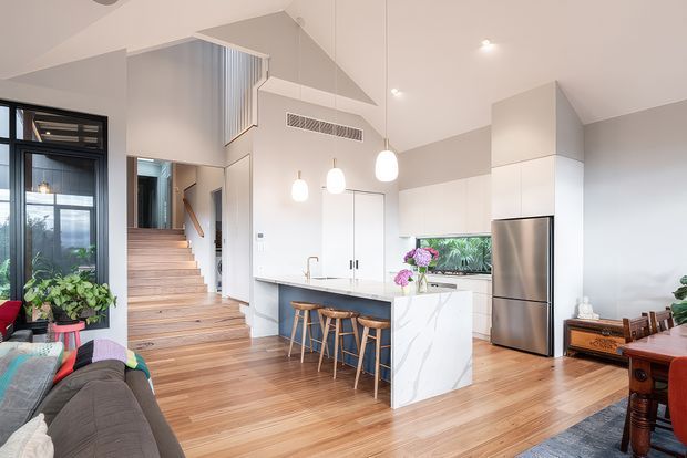Bright open-plan contemporary living, dining and kitchen area with warm timber flooring, white cabinetry, a marble-look island bench, stainless-steel refrigerator, pendant lights and timber staircase leading to an upper level. Large glazing and indoor plants add a light, airy feel.