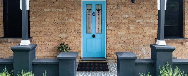 Front entrance facade of a traditional brick house, featuring a bright blue glazed-panel door, dark-painted window frames and rendered porch columns with decorative bases. Patterned tiled paving and low front boundary walls frame the entry, with small plants at the edges.