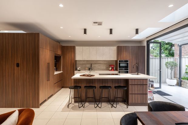 Contemporary open-plan kitchen with a large island and four bar stools. The kitchen features rich vertical timber cabinetry, a pale stone island benchtop, integrated stainless-steel appliances, a tiled floor, recessed ceiling lighting, and generous glazed openings leading to an outdoor courtyard or terrace.