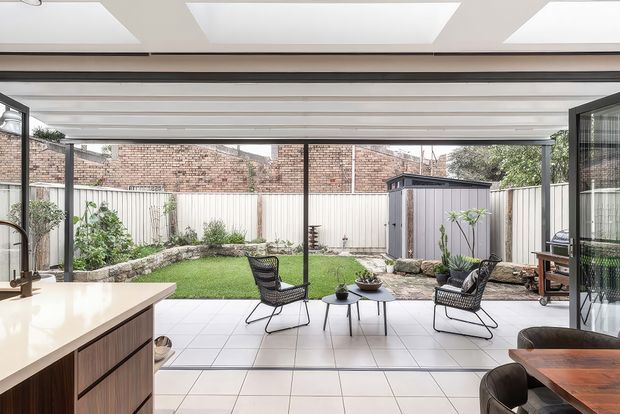Bright contemporary indoor-outdoor living space with a fully glazed, wide-opening facade overlooking a compact landscaped courtyard. The interior includes a pale tiled floor, kitchen island with timber cabinetry, and a dining area, while the covered outdoor zone has lounge chairs and a small table. Exposed brick boundary walls, a shed, raised garden bed and large roof skylights are visible.