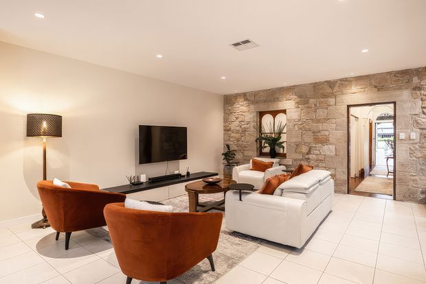 Bright, spacious living room with a contemporary seating arrangement, including a white sofa and rust-orange lounge chairs around a low coffee table. A wall-mounted television and slim media console sit against a plain painted wall, while a prominent exposed stone feature wall incorporates artwork, a doorway, and an arched passage beyond. Large light floor tiles, recessed ceiling lights, and warm neutral finishes create a refined, inviting interior.