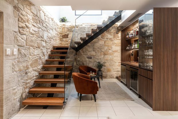 A refined interior stair hall with exposed rough stone walls, a floating timber staircase with a clear glass balustrade and dark metal stringer, and a built-in dark timber bar or drinks cabinet with glass-fronted storage. Two rust-coloured lounge chairs sit beside a small round table on a tiled floor. A skylight brings natural light into the compact, multi-level space.