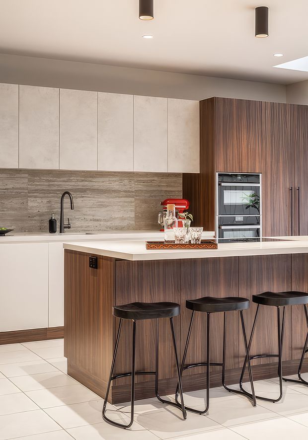 A contemporary kitchen with a large island and breakfast bar, featuring vertically grained dark timber cabinetry, pale stone or engineered-stone benchtops, a textured stone-look backsplash, integrated appliances, and sleek black bar stools. Recessed and pendant-style ceiling lighting create a warm, refined interior.
