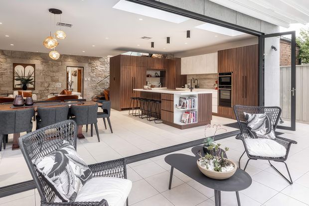 Open-plan contemporary kitchen, dining and living area with a large timber island and integrated cabinetry, white stone benchtops, a textured exposed-stone feature wall, and expansive glazed doors opening to an outdoor sitting area. The bright space includes a skylight, pendant lighting, bar seating, dining furniture and patterned lounge chairs.
