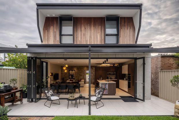 Contemporary two-storey residential extension viewed from the rear, with a covered outdoor entertaining area opening into an open-plan living, dining and kitchen space. The upper level features vertical timber cladding and dark-framed windows, while the ground floor has extensive retractable glazing and a tiled terrace with outdoor seating.
