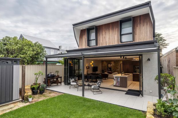 Exterior view of a contemporary two-storey home with a timber-clad upper level, dark metal roof and framing, and large glazed openings. The ground floor opens onto a paved patio and landscaped lawn, revealing an open-plan kitchen, dining and living area with warm timber cabinetry and a stone-textured feature wall. A covered outdoor entertaining area connects the interior to the garden.