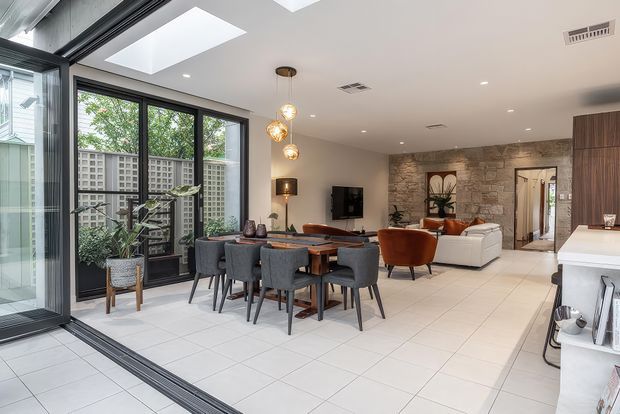 Open-plan contemporary living and dining area with large black-framed glazed doors, white tiled flooring, a timber dining table with upholstered chairs, and a lounge zone arranged around a television. A textured natural-stone feature wall and timber cabinetry add warmth, while roof skylights and extensive glazing provide abundant natural light.