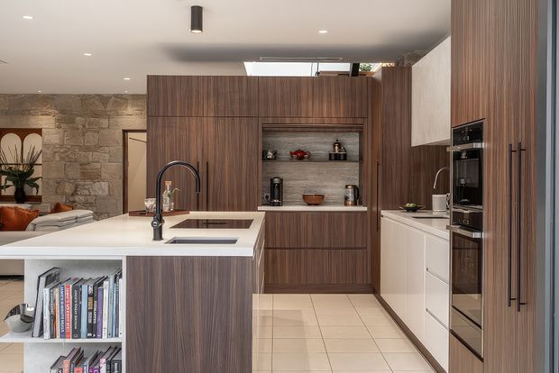 Contemporary kitchen with a large island featuring an integrated sink and dark gooseneck tap, warm vertical-grain timber cabinetry, white benchtops, and a built-in oven wall. Open shelving displays small appliances and ceramics, while a textured stone feature wall and glimpses of the adjoining living area add warmth and depth.