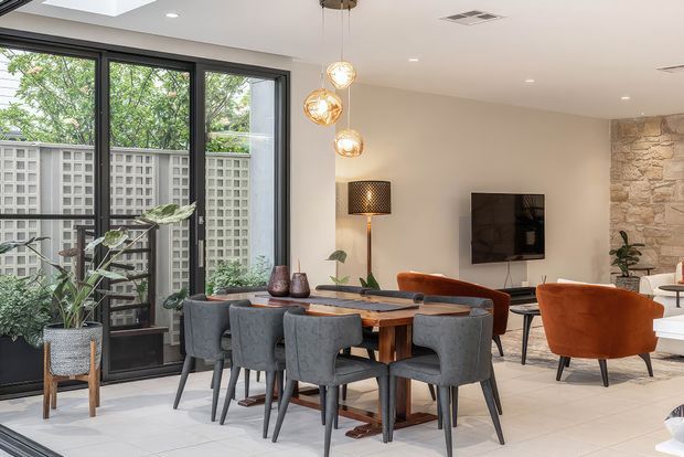A bright open-plan dining and living area with a large timber dining table surrounded by upholstered grey chairs, adjacent rust-orange lounge chairs, and a wall-mounted television. Floor-to-ceiling black-framed glazing opens to a leafy screened courtyard, while a natural stone feature wall, pendant lights, indoor plants, and pale tiled flooring add texture and warmth.