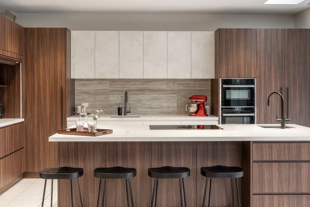 Contemporary galley-style kitchen with a large white island and four black bar stools, dark vertical wood-grain cabinetry, pale stone-look upper cabinets, a textured horizontal backsplash, integrated double ovens, and two black gooseneck faucets. A skylight or roof window brings in natural light.