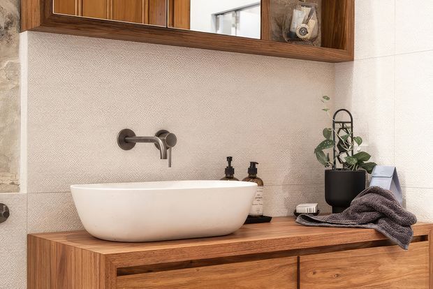 A warm, refined bathroom vanity with a white vessel basin set on a richly grained timber cabinet. The wall-mounted bronze-toned tapware, textured pale wall finish, timber-framed mirrored cabinet, toiletries, plant, and folded towel create a natural, contemporary setting.