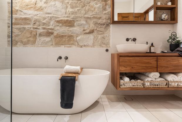 A refined contemporary bathroom featuring a freestanding white bathtub, walk-in glass shower screen, wall-mounted white basin, and timber vanity with open woven storage baskets. The room combines large pale floor tiles, smooth light wall tiles, a warm rough-stone feature wall, and dark metal tapware, with a timber-framed mirrored cabinet above the vanity.