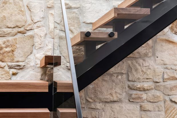 Close-up architectural view of a contemporary staircase with dark structural steel stringers, warm timber treads, and a slim glass balustrade set against an exposed rough stone masonry wall.