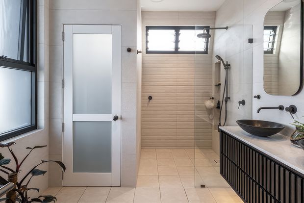 Bright contemporary bathroom featuring a walk-in shower with a timber-look tiled feature wall, pale large-format floor and wall tiles, black-framed louvre windows, a frosted-glass internal door, and a floating vanity with black vertical slatted cabinetry, a white benchtop, countertop basin, and large rounded mirror. Black tapware and shower fittings provide contrast.