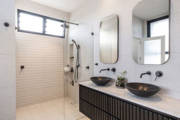 Bright contemporary bathroom with a walk-in glass shower, twin black vessel basins, wall-mounted black tapware, rounded rectangular mirrors, and a long fluted dark timber vanity. Large pale tiles and clerestory windows create an airy, refined space.