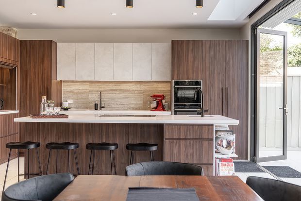 A contemporary open-plan kitchen and dining area with a large island and four black bar stools. The kitchen features dark timber-grain cabinetry, pale stone-look upper panels and benchtops, a textured timber-look splashback, integrated appliances, and a full-height glazed door opening to an outdoor area. A timber dining table and upholstered dining chairs occupy the foreground.