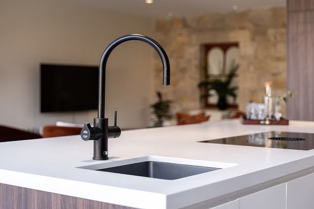 Close-up view of a contemporary kitchen island with a white solid-surface benchtop, integrated rectangular sink, and tall matte-black gooseneck mixer tap. Dark wood-grain cabinetry sits below the island, while a living area with a wall-mounted television and stone feature wall is softly visible in the background.