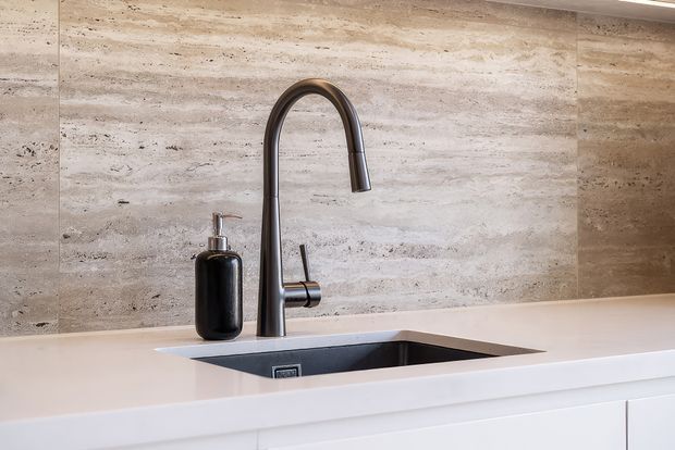 Close-up of a refined kitchen sink area with a matte dark arched gooseneck tap, integrated dark undermount sink, pale engineered-stone benchtop, and horizontally laid beige-grey natural stone splashback. Minimal fixed cabinetry is visible below.
