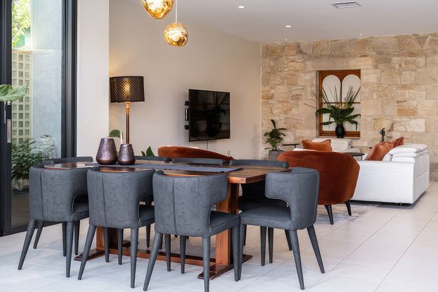 Open-plan dining and living area with a large dark timber dining table surrounded by upholstered charcoal dining chairs. The space features pale tiled flooring, warm neutral walls, a textured natural-stone feature wall with a recessed decorative niche, contemporary pendant lights, and a comfortable lounge arrangement in rust, tan, and white upholstery. Large glazing and indoor plants add natural light and greenery.