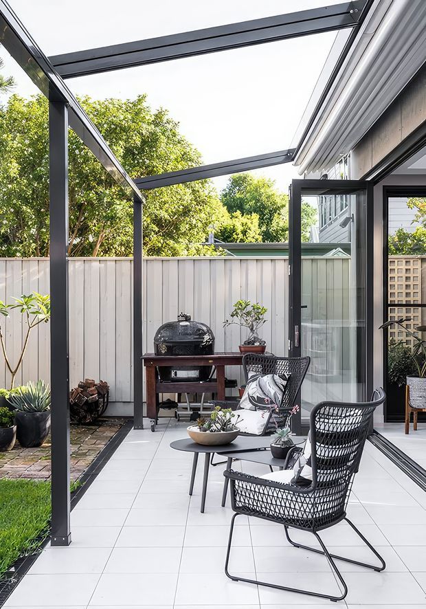 A bright outdoor patio or terrace with large pale tiled flooring, black-framed glazed doors, and a slim black steel pergola structure overhead. The space is furnished with woven black lounge chairs and a small coffee table, while a dark barbecue sits against a light vertical-panel fence. Potted plants, firewood storage, and a trellis add greenery and texture.