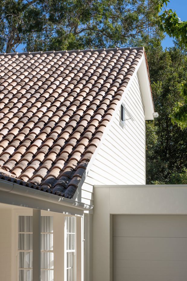 Close-up exterior view of a light-coloured house featuring a prominent terracotta barrel-tile pitched roof, white horizontal weatherboard cladding, white trim and gutters, multi-pane glazed doors, and an attached white garage. Mature trees surround the building.