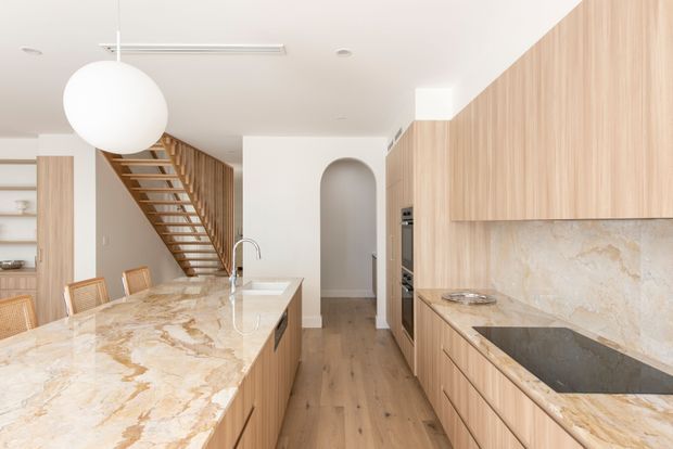 Bright contemporary kitchen with a large veined stone island, timber cabinetry, matching stone splashback and benchtops, and warm timber flooring. A sculptural white globe pendant hangs above the island, while a glazed balustrade staircase and arched wall niche are visible in the background.
