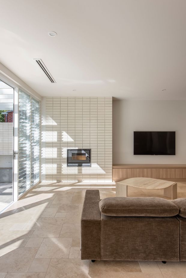 Bright contemporary living room with a large glazed opening and louvred window wall on the left, a tiled feature wall containing a built-in fireplace, and a wall-mounted television above fixed timber joinery. A brown upholstered sofa and light timber coffee table face the television. Large pale stone-look floor tiles and abundant sunlight create an airy, refined interior.