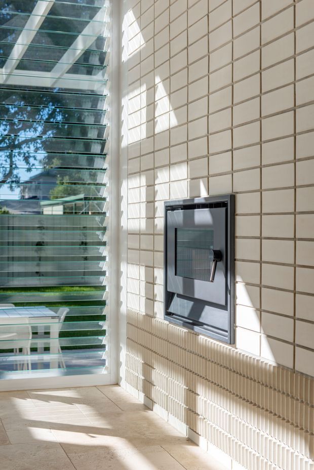 Sunlit interior with a large louvred glass window wall on the left and a cream tiled feature wall on the right. The wall incorporates a dark built-in fireplace or heating unit, while the pale stone or tile floor and strong geometric shadows create a bright, contemporary architectural space.